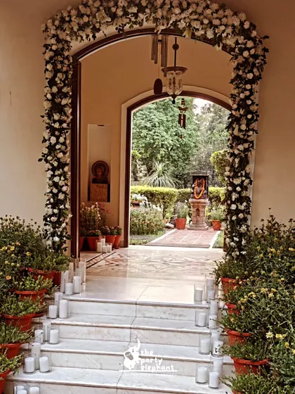 The wedding entrance in daylight, showing the grand floral arch and the arrangement of potted plants and unlit candles in preparation for the evening ceremony.
