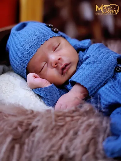 A close-up of a sleeping newborn in a cozy blue knit outfit. The texture of the fabric and the soft fur create a warm and comforting setting for the baby.