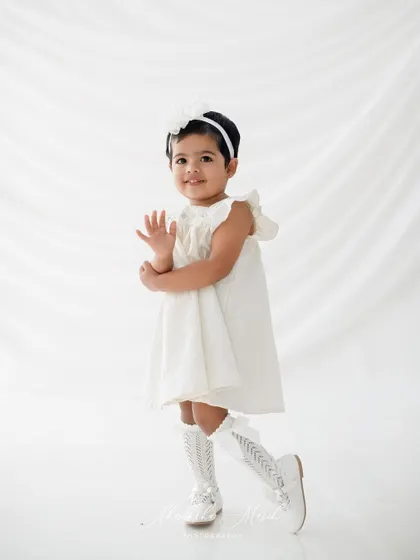 A playful wave from the birthday girl. Dressed in a classic white dress, she is all smiles and charm for her second birthday photoshoot.