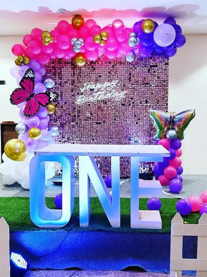 A stylish first birthday setup with a butterfly theme. The decor features a "ONE" table, a pink sequin wall, and "BABY" blocks, all on a turf-covered stage.