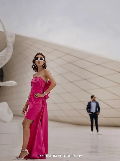 The bride strikes a pose in her vibrant pink dress in front of a modern sculpture, with the groom in the background.