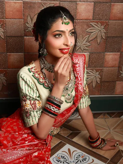 A seated portrait of a model, her smile lighting up the photo. The traditional red and white saree and heavy jewelry create a classic bridal look, captured in a relaxed and happy pose.