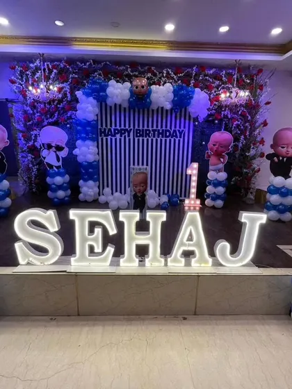 A Boss Baby stage decoration for a first birthday. This setup includes a striped backdrop, a flower wall, and large illuminated letters spelling the name 'SEHAJ'.