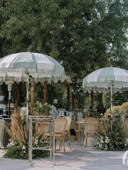 An outdoor seating area designed for a classic ivory and green themed wedding. Fringed umbrellas and comfortable seating are surrounded by arrangements of pampas grass and white florals.