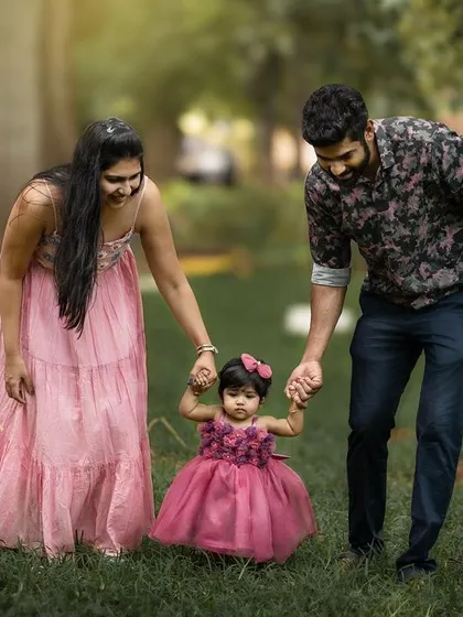 A beautiful outdoor family portrait, with parents helping their little girl take her first steps on the grass. A perfect memory of a precious milestone.