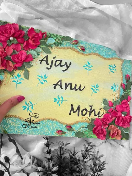 A rectangular nameboard for "Ajay, Anu, Mohi," featuring deep red roses on a neutral yellow base with turquoise accents. The black and white background makes the colors pop.