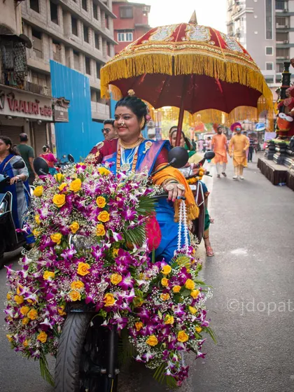 A woman rides a scooter adorned with a massive arrangement of flowers during the Gudi Padwa festival, with a traditional umbrella held behind her.