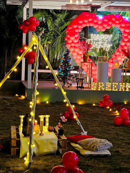 A wide view of a romantic garden party, featuring a large red balloon heart, a lit-up teepee, and low picnic seating.
