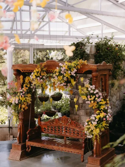 A traditional wooden swing, or 'jhoola', beautifully decorated for the Haldi ceremony. It's adorned with cascading arrangements of yellow and pink flowers, creating a perfect focal point within the greenhouse.