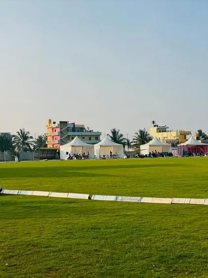 Players and spectators can be seen relaxing by the boundary during a break in play, with the tournament tents visible in the background.