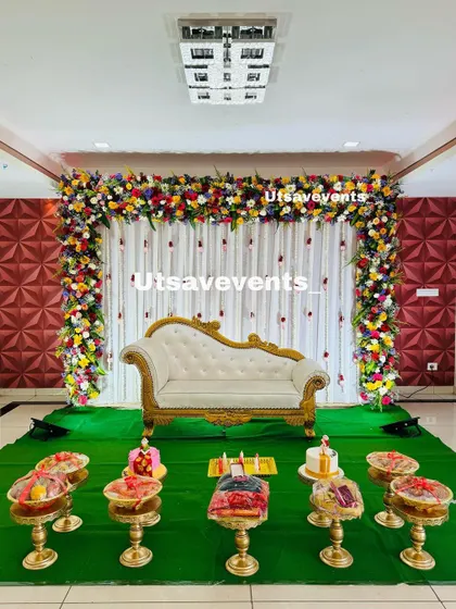 A simple and elegant baby shower stage with a multi-colored floral arch over a white curtain backdrop. The setup includes a white divan and brass stands for offerings.