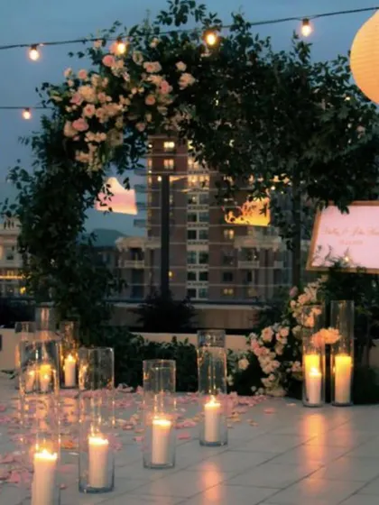 A soft, romantic proposal setting with a circular floral arch against the city skyline. The aisle is lined with candles in tall glass holders, creating an elegant and serene atmosphere for a sunset proposal.