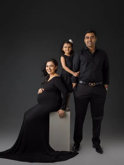 A stylish and modern family portrait. The expecting mother is seated while her partner and daughter stand beside her, all dressed in elegant black, creating a powerful and chic image.
