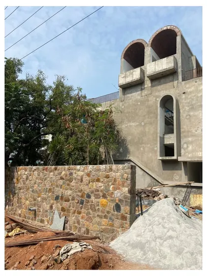 The exterior of the Brick Vault house during construction, with the random rubble masonry wall in the foreground. The design retains the original lower floor while adding new volumes above, all compliant with Vastu principles.