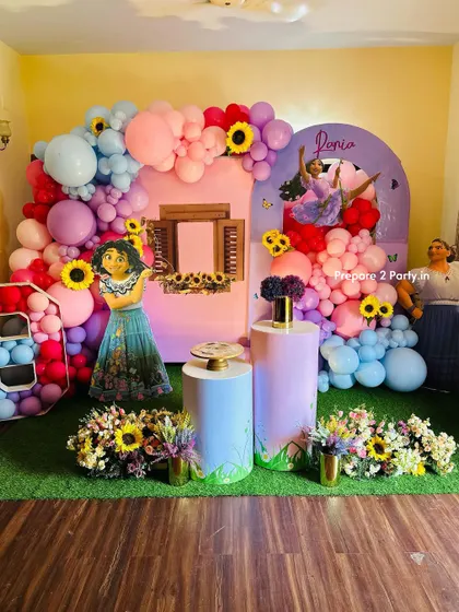 A magical Encanto themed birthday party, complete with a backdrop of the Casita. The decoration features character cutouts of Mirabel and Luisa, vibrant balloon garlands, and sunflower accents to capture the movie's charm.