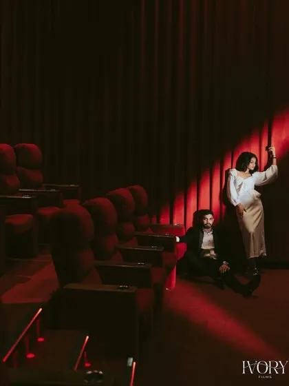 An artistic shot using a single spotlight against the rich red curtain of the theatre. The composition is creative and moody, capturing a quiet, cinematic moment between the couple.