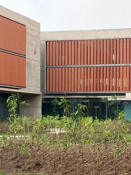 The JSW Steel office nearing completion, with newly planted landscaping in the foreground. The terracotta-colored fins and raw concrete facade are key elements of its climate-responsive design.