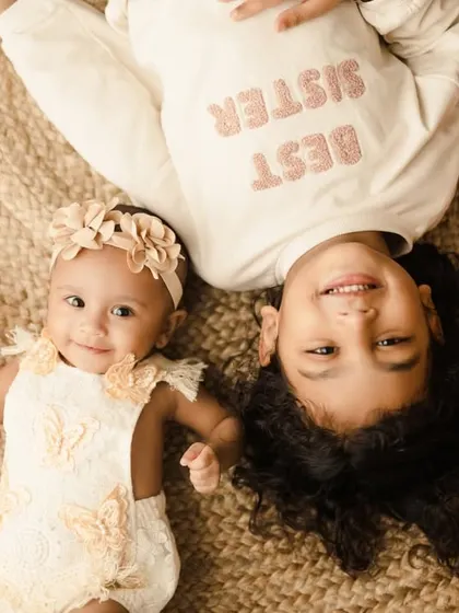 An adorable overhead shot of two sisters lying on a jute rug. The older sister's "Best Sister" shirt is a cute detail in this playful sibling portrait.