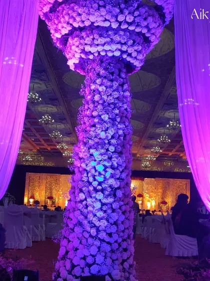 The purple lighting enhances the texture and grandeur of this towering floral column, making it a focal point of the event's design.