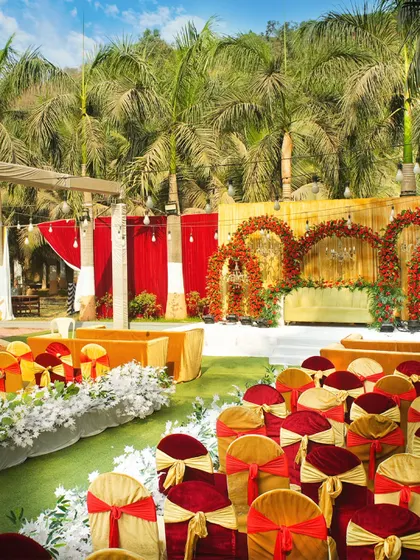 An outdoor wedding ceremony setup with a vibrant red and gold theme. The seating is arranged to give every guest a perfect view of the beautiful floral mandap.