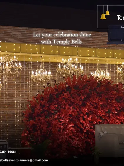 A close up of a luxurious reception stage detail. The backdrop features a curtain of warm fairy lights and mini chandeliers behind a large arrangement of red flowers, creating a rich and opulent look.