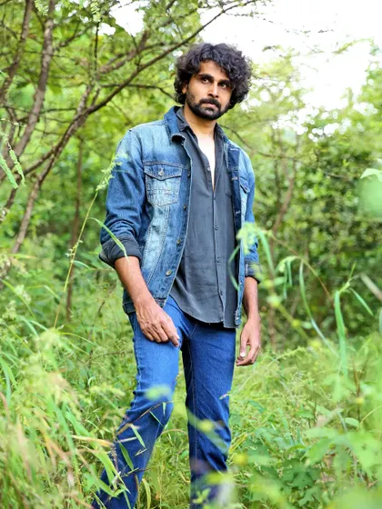 A full-body portrait for a men's fashion portfolio, set against a lush green backdrop. The denim-on-denim outfit stands out while the natural environment adds a rustic, authentic feel to the photograph.
