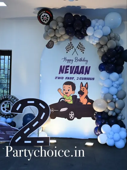 A "Two Fast, 2 Curious" car-themed birthday party. This setup includes a large number '2' cutout designed as a racetrack, a custom backdrop, and a balloon arch in shades of blue, black, and grey.
