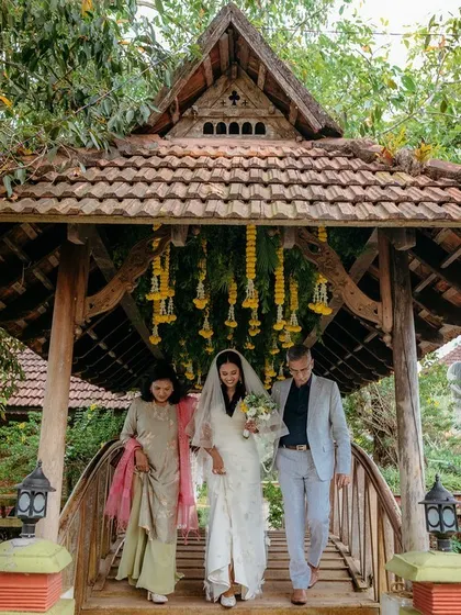 A bride is escorted over a charming wooden bridge at a resort in Kerala, showcasing a beautiful destination wedding with cross-cultural elements.