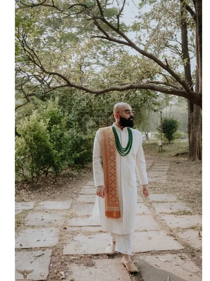 A solo portrait of the groom, looking thoughtful and elegant in his white sherwani and green necklace, set against a natural, park-like background.