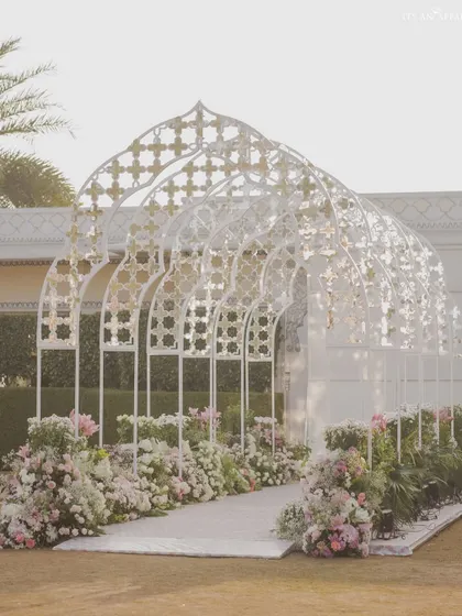 The grand entrance tunnel for a Mughal-themed wedding. The intricate white jali arches, lined with pastel flowers, create a breathtaking walkway for guests arriving at the ceremony.