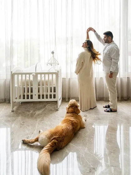 A beautiful, wide shot of an expecting couple dancing by a window and a crib, while their golden retriever lies on the floor watching them.