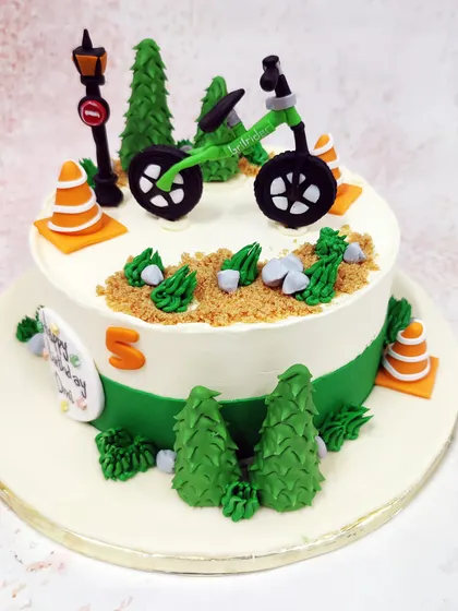 We set the scene for an outdoor adventure with this cycle theme cake. A green bicycle is parked on a dirt path surrounded by pine trees and traffic cones, creating a fun and detailed landscape for a young cyclist's birthday.