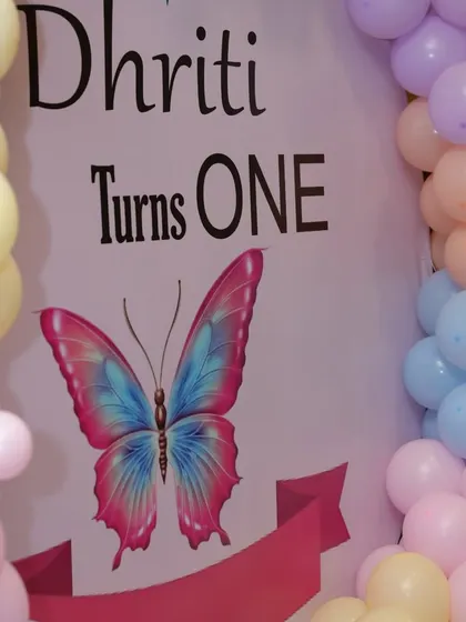 A close-up of the "Dhruti Turns ONE" backdrop from a butterfly-themed party. The pastel colors are soft and perfect for a first birthday.