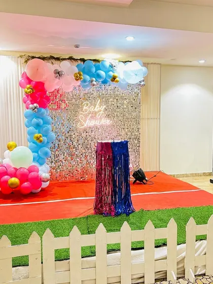 A gender reveal baby shower setup with a silver shimmer wall. The stage is decorated with pink and blue elements, including balloon columns topped with baby foil balloons.