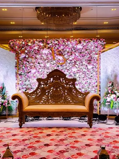 An elegant reception stage featuring a carved wooden sofa set against a wall of pink and white flowers. The patterned carpet and panelled backdrops add a layer of texture and sophistication.