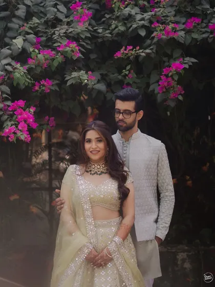 A classic portrait of a couple at their Mehendi ceremony, framed by beautiful pink bougainvillea. Their elegant, coordinated outfits and the lush floral background create a timeless and romantic image.