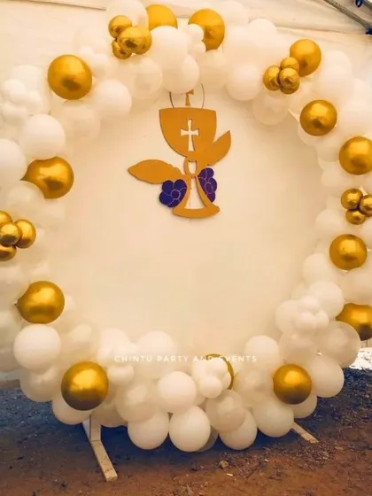 An outdoor baptism ceremony decoration with a white and gold balloon ring. A cutout of the holy chalice is placed in the center.