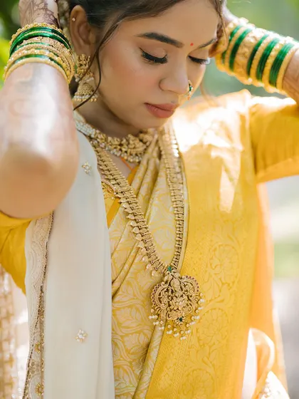A close-up portrait of the bride adjusting her intricate gold necklace. The focus is on her delicate features, traditional jewelry, and the rich texture of her silk saree.