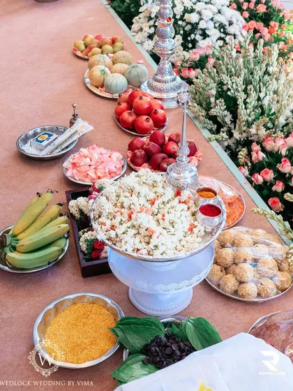 A beautiful display of offerings for a traditional South Indian ceremony. The arrangement includes fresh fruits, flowers, sweets, and other symbolic items, set against a backdrop of lush pink and white florals.