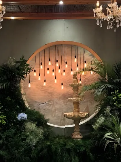 Creating moments of quiet romance is my passion. This enchanting photo corner, designed for an engagement party, features a classic fountain set against a warm, glowing backdrop and surrounded by lush, wild foliage and hanging Edison bulbs.