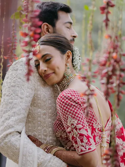 A close-up, intimate portrait of a couple embracing, with the bride resting her head on the groom's shoulder amidst a backdrop of red flowers.