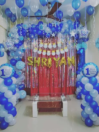 A "First Birthday Boy" themed decoration in shades of blue and white. This full room setup includes a balloon arch, shimmer backdrop, and multiple themed foil balloons to celebrate the special milestone.