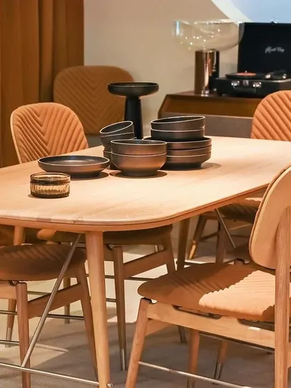 A closer view of the Volta Oak Dining Set. The smooth, rounded edges of the table and the comfortable, upholstered chairs create an inviting atmosphere for any meal. The record player in the background hints at the collection's musical inspiration.