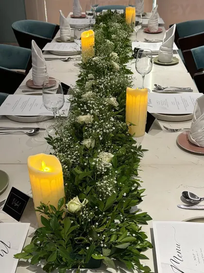 This is an elegant dining table featuring a lush centerpiece made of fresh greenery and baby's breath. The table is set with custom menus, place cards, and tall electronic pillar candles for safe, ambient lighting.