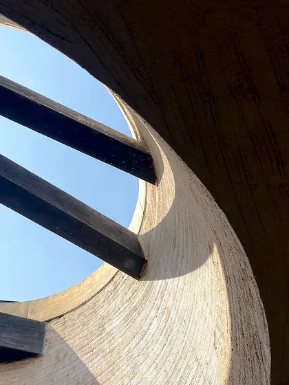A close-up of the skylight detail, where the raw texture of the plasterwork catches the sunlight. The wooden rafters cut across the circle of blue sky, creating a strong graphic composition.