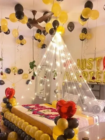 A "Just Married" bedroom decoration with a black and yellow theme. The setup includes a light-up canopy, balloons on the bed and ceiling, and scattered rose petals.