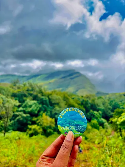 The Kudremukha trek badge held up against a backdrop of the peak on a beautiful, cloudy day.