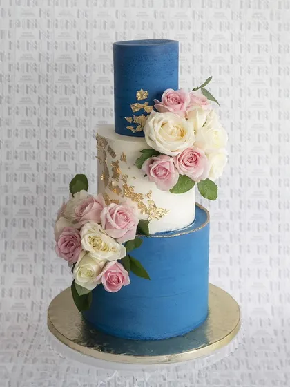 A stunning three-tier wedding cake combining bold blue and classic white. The middle tier is accented with gold leaf and the entire cake is adorned with beautiful pink and white roses.