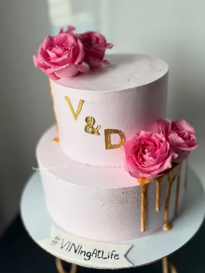 A lovely two-tiered cake for a special celebration, featuring soft pink frosting, a gold drip, and the couple's initials. It's decorated with fresh pink roses to complete the romantic look.