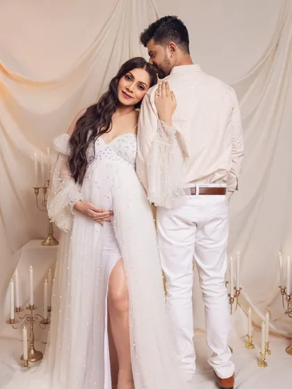 An angelic and romantic maternity setup. The mother-to-be, in a stunning white gown with pearl details, leans against her partner, surrounded by the soft glow of candles for a truly magical portrait.